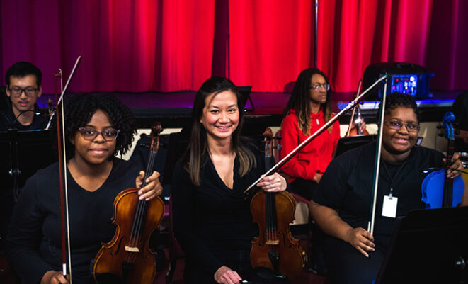 SLSO musician Jessica Cheng Hellwege sitting between two Jennings violin students in orchestra pit