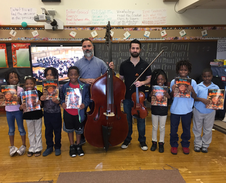 SLSO musicians David DeRiso and Chris Tantillo holding string instruments standing in the center of seven elementary age students holding books