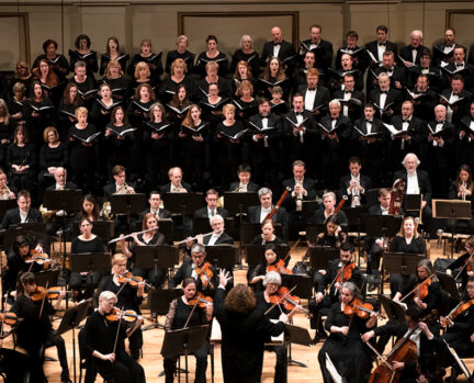 Chorus members singing on stage with the orchestra playing